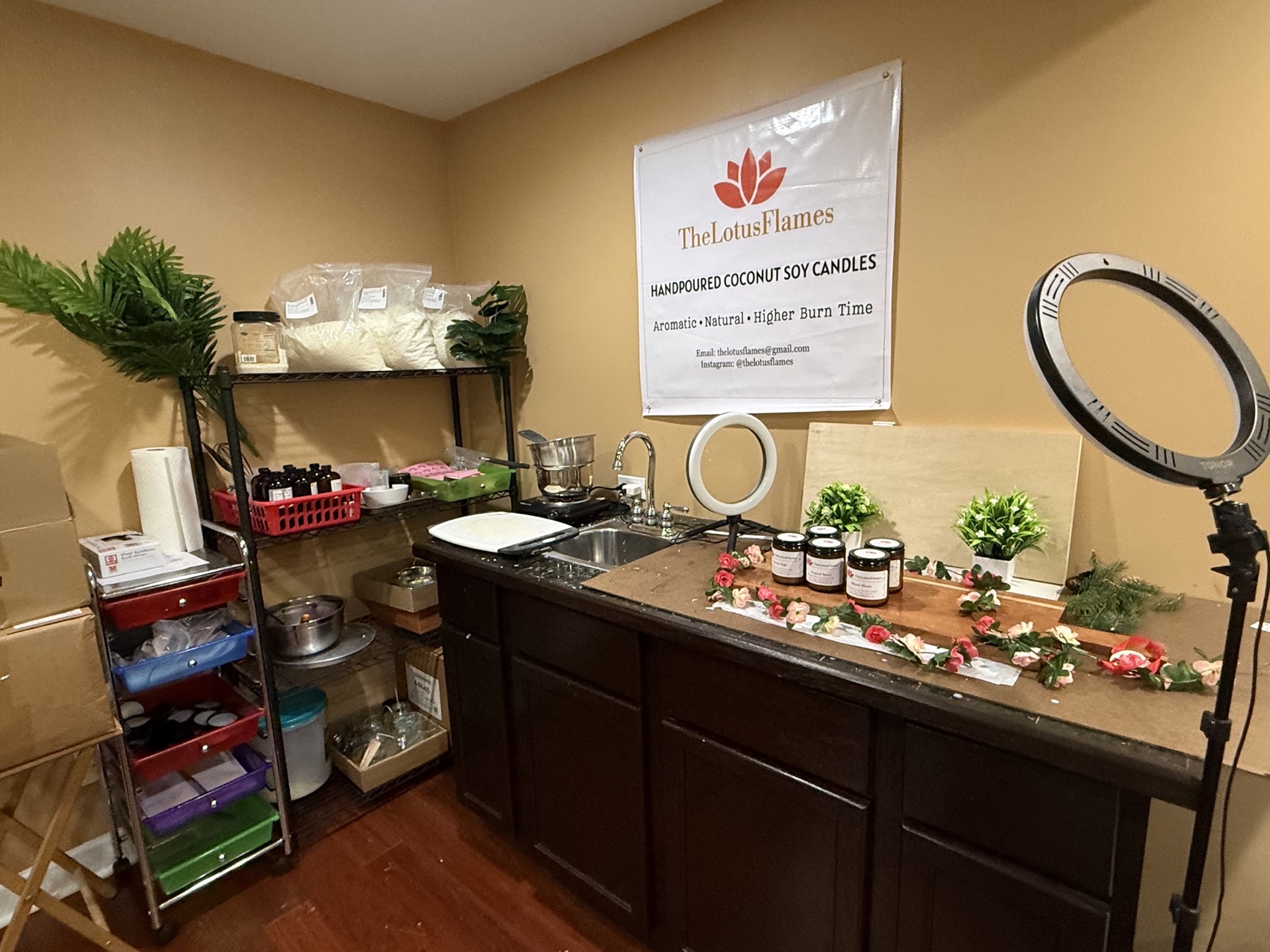 The TheLotusFlames candle studio — coconut soy wax, moulds and supplies on wire shelving, with hand-poured candles and flowers set up under ring lights beneath the TheLotusFlames banner.